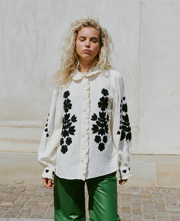 Woman wearing a Sissel Edelbo creamy white blouse with black floral patterns against a light-colored wall.