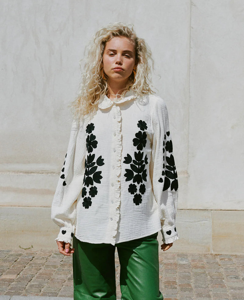 Woman wearing a Sissel Edelbo creamy white blouse with black floral patterns against a light-colored wall.
